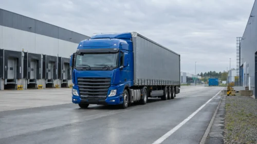 Niebieski ciągnik z naczepą na drodze wewnętrznej pomiędzy halami centrum logistycznego.